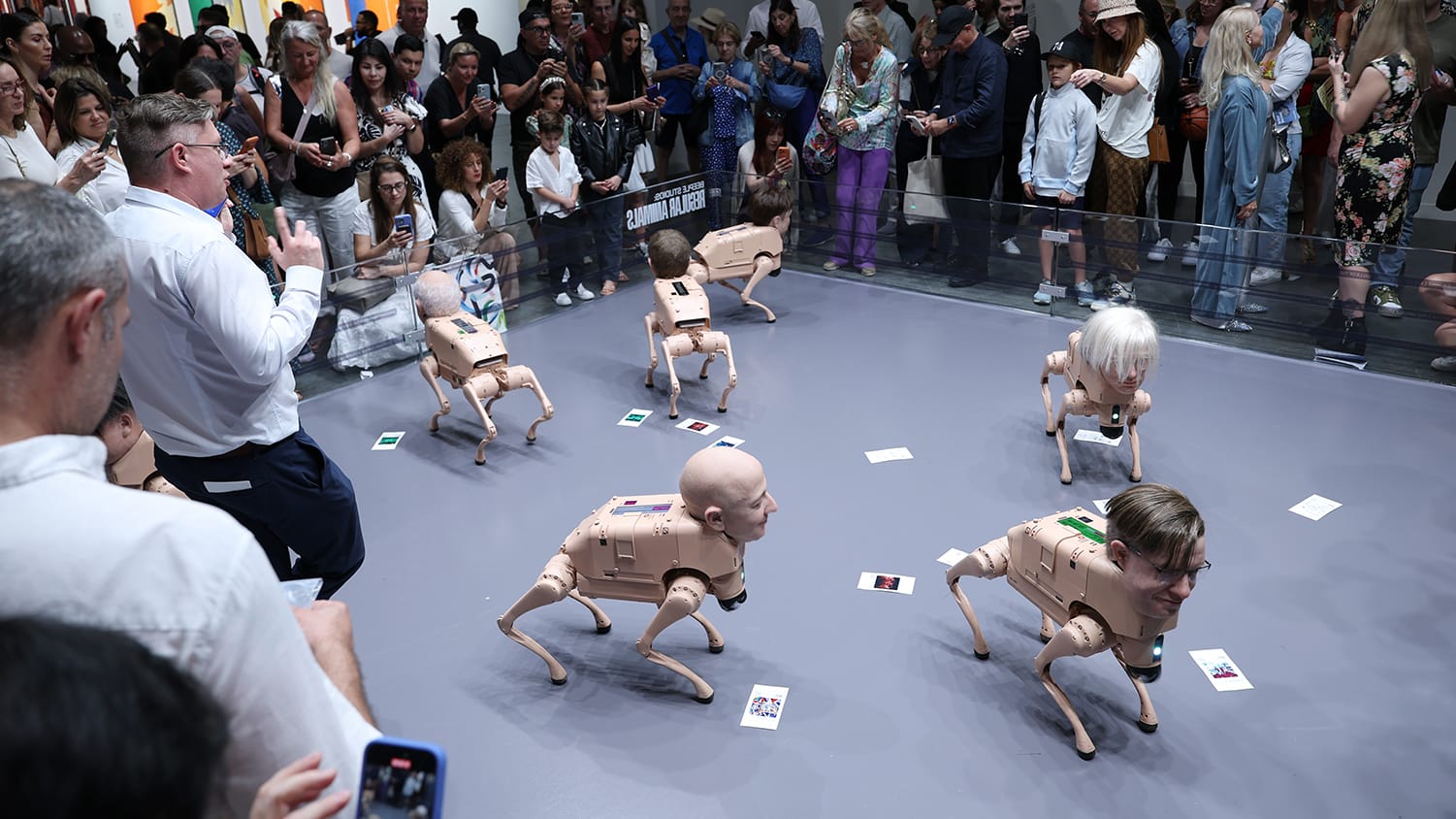 A crowd of people gather around an art exhibit of robot dogs with human heads.